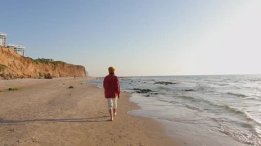 Sahilde yürüyen gömlekli bir kadın ve arkasında güzel bir günbatımı var. Yaz tatilindeki turist. Gülümseyen neşeli bayan sahilde eğleniyor ve güzel doğanın tadını çıkarıyor.. 