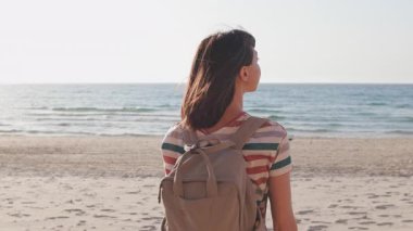 Sırt çantalı genç bir kadın gün batımında sahilde duruyor, doğanın huzur ve güzelliğinin tadını çıkarıyor. Kız kameraya bakar ve gülümser..