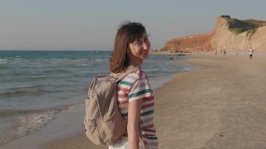 Sırt çantalı genç bir kadın gün batımında sahilde yürüyor. Doğanın huzur ve güzelliğinin tadını çıkarıyor. Kız kameraya bakıp gülümsüyor..