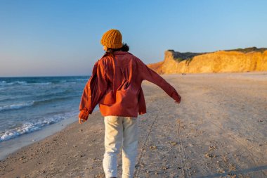 Sahilde yürüyen gömlekli bir kadın ve arkasında güzel bir günbatımı var. Yaz tatilindeki turist. Gülümseyen neşeli bayan sahilde eğleniyor ve güzel doğanın tadını çıkarıyor..