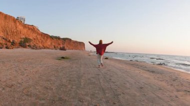 Sahilde gün batımını seyreden tişörtlü ve şapkalı neşeli bir kadın. Arkadan bak. Tatilde bir turist. Havalı kız sahilde eğleniyor. Güzel doğanın tadını çıkarıyor..