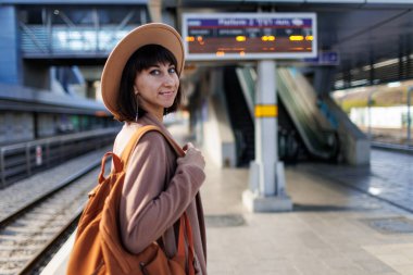 İstasyonda sırt çantalı bir kız. Tren istasyonundaki güzel ve genç bir kızın portresi. Paltolu ve şapkalı genç bir kadın istasyon platformunda duruyor. Maceralar ve seyahat.