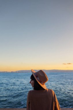 Paltolu ve şapkalı çekici genç bayan gün batımını seyrediyor. Kız denize bakmaktadır. sakinlik ve sükunet.