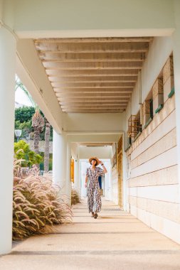 Şapka takan güzel bir bayanın tam boy fotoğrafı. Sokakta yürümek, güneşli hava, hafta sonu. Elbiseli ve şapkalı genç bir kız sokakta yürüyor..