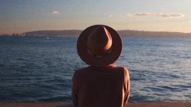 Gün batımında güzel deniz manzarasının tadını çıkaran genç bir kadın ceketli ve şapkalı çekici bir kadın gün batımını seyrediyor. Denize bakan kız. Sakin ve huzurlu.