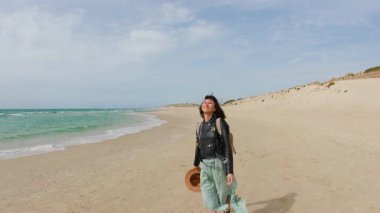 Boş kumsalda yürüyen mutlu genç kız, deniz arka planında esintiden hoşlanan kaygısız kadın. Seyahat, tatil, açık hava eğlencesi..