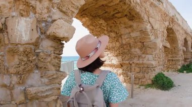 Şapka takan ve sırt çantası taşıyan kadın gezgin antik bir su kemerinin yanında yürüyor ve tarihi Caesarea, İsrail 'i keşfediyor..
