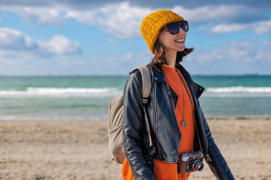 Soğuk havada kumsalda sırt çantalı bir kadın. Güneşli bir günde deniz kenarında yürüyüş yapan şık bir kadın. Sırt çantalı kız..