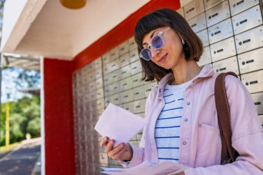 Bir kadın posta kutusunun önünde beklerken zarf ve mektupları inceliyor. Alıcı bir banka hesabından tebligat alır. Zarf tutan bir kız.