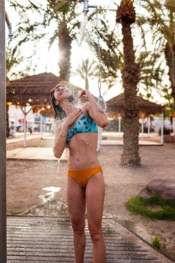 Genç kız tropikal bir duş alıyor. Kumsalda duş al. Kadın vücudunu yıkıyor. Mayo giymiş bir kadının duş alırken görüntüsü. Plajda bikinili bir kız. Yaz havası.