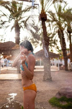 Genç kız tropikal bir duş alıyor. Kumsalda duş al. Kadın vücudunu yıkıyor. Mayo giymiş bir kadının duş alırken görüntüsü. Plajda bikinili bir kız. Yaz havası.