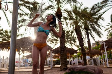 Genç kız tropikal bir duş alıyor. Kumsalda duş al. Kadın vücudunu yıkıyor. Mayo giymiş bir kadının duş alırken görüntüsü. Plajda bikinili bir kız. Yaz havası.
