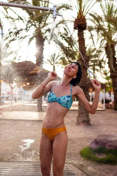 Mayo giymiş ince bir kadın gün batımında duş alıyor. Kız sahilde duş alıyor, deniz kenarında. Yaz havası. Mayolu kız..