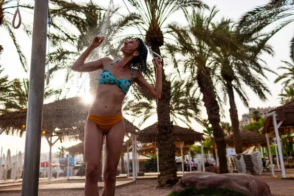 Genç kız tropikal bir duş alıyor. Kumsalda duş al. Kadın vücudunu yıkıyor. Mayo giymiş bir kadının duş alırken görüntüsü. Plajda bikinili bir kız. Yaz havası.