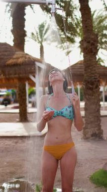 Dikey çekim. Genç kız tropikal bir duş alıyor. Kumsalda duş al. Kadın vücudunu yıkıyor. Mayo giymiş bir kadının görüntüsü...