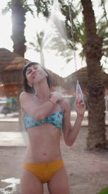 Dikey çekim. Yaz havası. Mayo giymiş esmer, beyaz bir kadın lüks bir otelde serinletici bir tropikal duş alıyor.