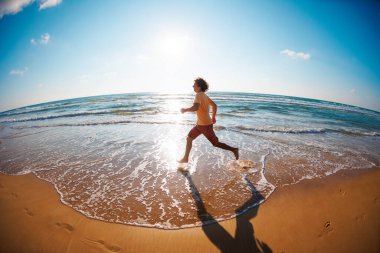 Güneşli bir yaz gününde, spor kıyafetli bir adamın spor salonunda kumlu sahil boyunca tek başına koşuşunun yan görüntüsü. Kumda koşarken.