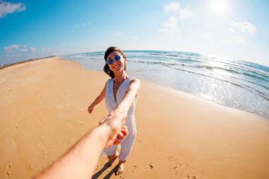 Beni takip edin. Genç ve mutlu bir kadın erkeklerin elini çekiyor. Elbise giymiş güzel bir kız sahilde yürüyor, bir erkeğin elini tutuyor, romantik bir yürüyüş. Sevgililer Günü. Sahil boyunca yürü..