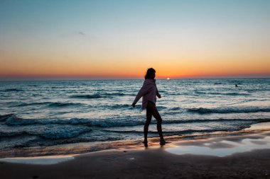 Gün batımında sahilde yürüyüş yapan bir kız. Gün batımına bakan bir kadının silueti. Yaz havası. Sahildeki kız..