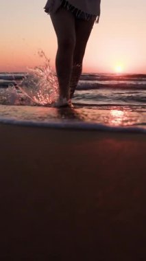 Dikey çekim. Gün batımında sahilde yürüyüş yapmaktan hoşlanan iri yarı genç bir kadın. Deniz kıyısında kumsalda yalınayak yürümek. Yaz