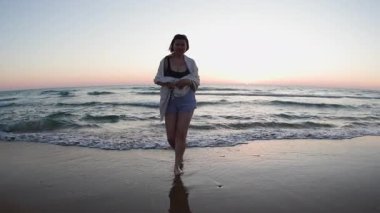 Gün batımında sahilde yürüyüş yapan iri yarı genç bir kadın. Sahilde çıplak ayakla yürüyen kadın ayaklarının yavaş hareketi.