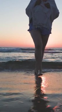 Dikey çekim. Gün batımında sahilde yürüyüş yapmaktan hoşlanan iri yarı genç bir kadın. Deniz kıyısında kumsalda yalınayak yürümek. Yaz