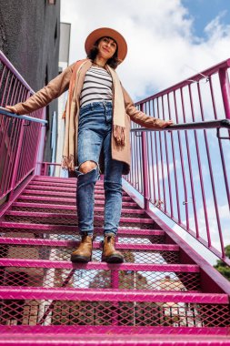 Şapkalı şık bir kız şehrin arka planına karşı korkulukların yanında poz veriyor. sokak tarzı ve moda.