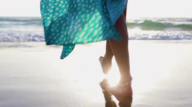 Gün batımında güzel, dalgalı elbiseli bir kadın. Yaz havası. Altın gün batımında sahilde çıplak ayakla yürüyen kadın ayaklarının yavaş hareketi.