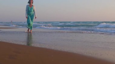 Romantik bir ruh hali. İnce elbiseli kadın kumlu sahilde dalgalar boyunca yavaşça yürüyor ve anın tadını çıkarıyor. Güzel kadın.