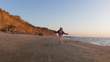 Boş sahilde koşan ve özgürlük hissiyle kollarını yanlara kaldıran genç bir kadın görüntüsü. Ön görünüm
