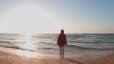 Okyanus sahilinde yürüyen bir kadın hayatının tadını çıkarıyor. Tişört ve şapka giymiş, kumlu yolda yürüyen mutlu kadın gezgin.