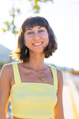 Havalı yaz kıyafetleri içinde, yüzü piercingli, gülümseyen genç bir esmer. Güneşli bir günde dışarıda poz veren baştan çıkarıcı, kaygısız kadın. Açık havada pozitif model. Yaz havası.