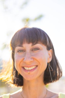 Havalı yaz kıyafetleri içinde, yüzü piercingli, gülümseyen genç bir esmer. Güneşli bir günde dışarıda poz veren baştan çıkarıcı, kaygısız kadın. Açık havada pozitif model. Yaz havası.