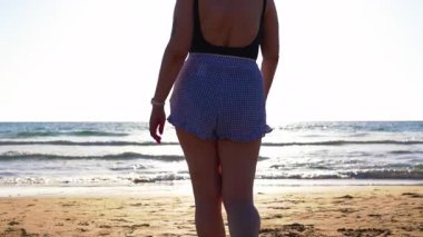 Gün batımında sahilde yürüyen bir kız. Ayrıca plajda ıslak kumda yürüyen ve vücudunun tadını çıkaran bir manken.