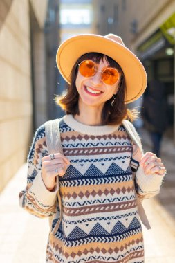 Şehirde sırt çantalı şık bir kızın portresi. Binaların arasında cadde boyunca yürüyen mutlu genç bir kadın. Sırt çantalı bir kadın gezgin şehri geziyor, kız şehirde dolaşıyor..