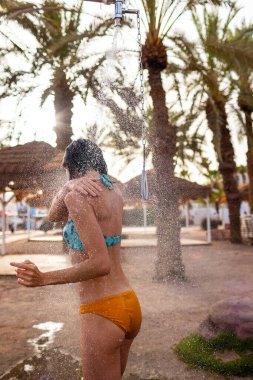 Mayo giymiş ince bir kadın gün batımında duş alıyor. Kız sahilde duş alıyor, deniz kenarında. Yaz havası. Mayolu kız..