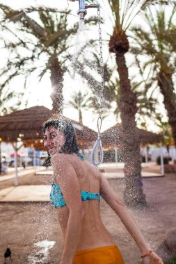 Genç kız tropikal bir duş alıyor. Kumsalda duş al. Kadın vücudunu yıkıyor. Mayo giymiş bir kadının duş alırken görüntüsü. Plajda bikinili bir kız. Yaz havası.