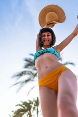 Tropikal plajda bikinili ve şapkalı güzel bir kadın. Yaz havası. Mutlu gezgin, tropik sahilde tatilin tadını çıkarıyor..