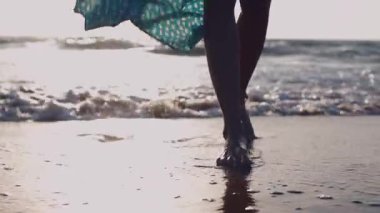 Gün batımında güzel, dalgalı elbiseli bir kadın. Altın gün batımına karşı sahilde çıplak ayakla yürüyen bir kadın. Yazın turist