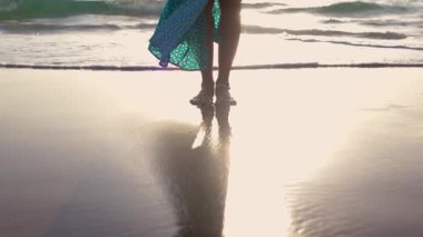 Dalgalı elbiseli kadın sahilde altın gün batımına ve okyanus dalgalarına karşı duruyor. Bacaklar yakın plan. Çıplak ayaklı bir kadın. Turist