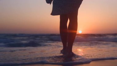 Gün batımında okyanus sahilinde yürüyen bir kadının arka görüntüsü. İnce kadın bacakları ıslak kuma basar, ayak izleri bırakır, ağır çekimde.