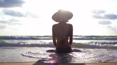 Yaz akşamları kumlu sahilde meditasyon yapan ince bir kadın. Kadın, okyanus dalgalarının yanındaki kumların üzerinde lotus pozisyonunda oturuyor.