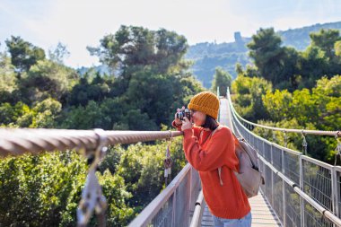 Şapkalı ve sırt çantalı şık genç bir kadın güneşli bir havada kamerayla fotoğraf çekiyor. Seyahat ve fotoğrafçılık. Yolculuğunun tadını çıkaran mutlu bir bayan turist. Sırt çantalı kız..