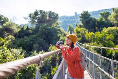 Kameralı ve sırt çantalı genç bir kadın asma köprü boyunca yürümekten hoşlanıyor. Yolculuğunun tadını çıkaran mutlu bir bayan turist. Sırt çantalı kız..