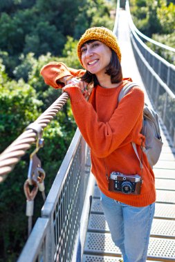 Seyahat ve fotoğrafçılık. Elinde kamera ve sırt çantası olan genç bir kadın asma köprü boyunca yürüyor. Seyahatinin tadını çıkaran mutlu bayan turist. Kameralı kız..