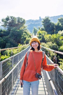 Kameralı ve sırt çantalı genç bir kadın asma köprü boyunca yürümekten hoşlanıyor. Yolculuğunun tadını çıkaran mutlu bir bayan turist. Sırt çantalı kız..