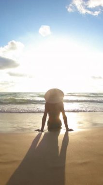 Dikey çekim. Deniz kıyısında oturan ve tatilinin tadını çıkaran bir kız. Arkadan bak. Deniz kenarında mayo giymiş, güneşlenen ince bir kız.