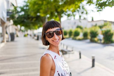 Genç güzel kız gülümser ve şehre bakar. Tarz sahibi genç bir kız sokakta yürüyor. Şehir tarzı, yaz trendi.