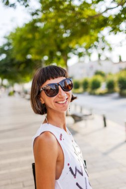 Güneşli bir caddede genç bir kızın portresi. Kız gülümsüyor ve kameraya bakıyor. Şehir tarzı, yaz trendi.