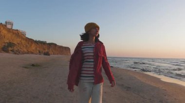 Genç bir kadın sahilde romantik tatilin tadını çıkarıyor. Gün batımının ve okyanus dalgalarının tadını çıkarıyor. Kahkaha ve mutluluk. Şık kız eğleniyor